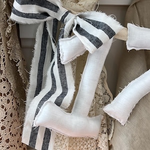 May include: A decorative fabric number seven in white linen with black and white striped accents. The number is embellished with a bow and is part of a home decor collection. The image shows a close-up of the number seven.