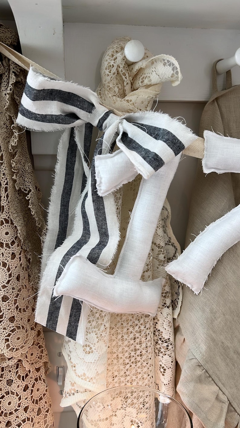 May include: A decorative fabric number seven in white linen with black and white striped accents. The number is embellished with a bow and is part of a home decor collection. The image shows a close-up of the number seven.