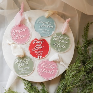 May include: Six round, clear acrylic ornaments with white calligraphy. The ornaments are decorated with pink, red, and green paint. The text on the ornaments reads: "Gracie Rose", "Connor Jordan", "Papa and Gigi Again in 2024", "Baby's 1st Christmas Olive Rhea 2022", "Big Sis Again Baby Webb Coming Summer 2024", and "Miller Nicole November".