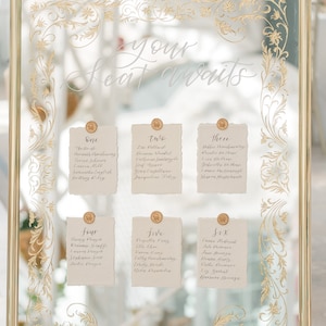 May include: A gold-framed mirror with a white inscription "your seat awaits" and six cards with names listed for a wedding reception seating chart.