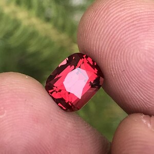 May include: A rectangular-cut, red gemstone with multiple facets. The gem is held between two fingers, with a blurred green background. The facets reflect light, creating a sparkling effect. The stone's colour is a deep, rich red.