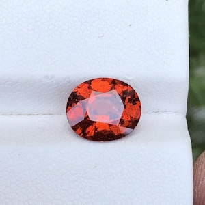 May include: A circular, cut gemstone in a vibrant red-orange shade. The gem is displayed against a white backdrop, highlighting its facets and internal light play. The gemstone, likely a garnet, exhibits a rich, fiery colour.