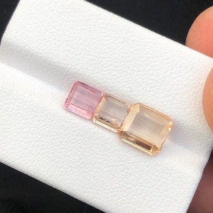 Puede incluir: Tres piedras preciosas facetadas, una rosa y dos melocotón, se muestran sobre una superficie blanca. Las piedras preciosas están talladas en forma rectangular.