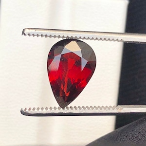 May include: A deep red pear-shaped gemstone held by tweezers against a white background.
