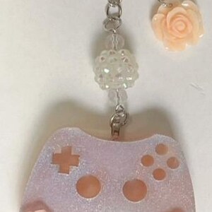 May include: A light purple glittery keychain with a video game controller design. The controller has a cross and button details, with a peach-coloured rose on each side. The keychain includes a chain, beads, and a peach-coloured rose charm.