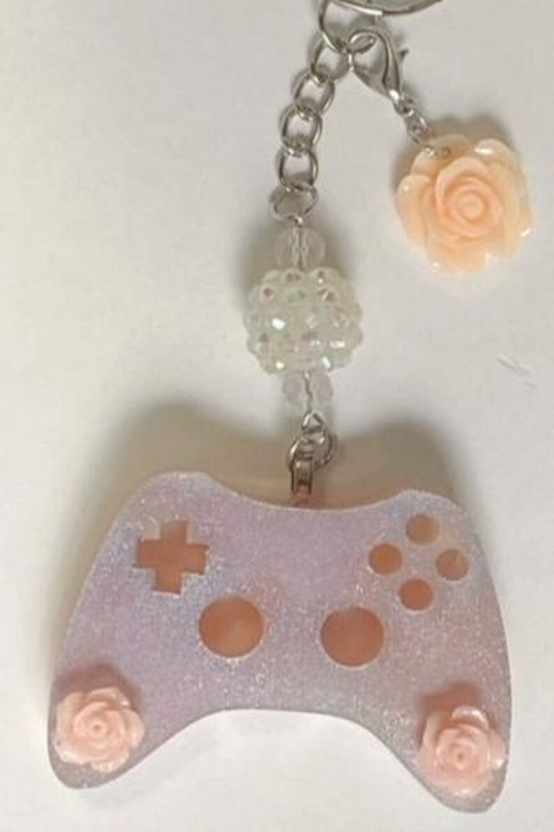 May include: A light purple glittery keychain featuring a video game controller design. The controller has a cross and button details, with a peach-colored rose on each side. The keychain includes a chain, beads, and a peach-colored rose charm.