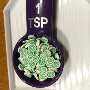 May include: A purple measuring spoon with a white label that reads "1 TSP". The spoon is filled with small, white and green swirl-shaped sprinkles.