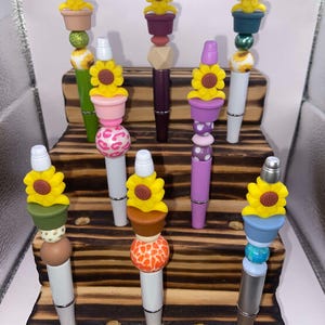 May include: A set of ten sunflower-themed pens with colorful beads and charms. The pens are displayed on a wooden stand with multiple slots.