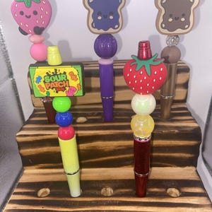 May include: Five colorful pens with different themed toppers displayed on a wooden stand. The toppers include a strawberry, a piece of toast with jelly, a piece of toast with chocolate spread, a strawberry, and a Sour Patch Kids candy.