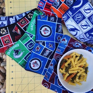 May include: A set of six colorful pot holders featuring various NHL team logos, including the Tampa Bay Lightning, Edmonton Oilers, Arizona Coyotes, Vancouver Canucks, Toronto Maple Leafs, and Montreal Canadiens. The pot holders are made of fabric and have a quilted design.