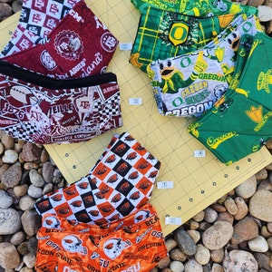 May include: Five different fabric face masks with various college logos and designs. The masks are laid out on a yellow cutting mat with a background of small rocks. The masks feature logos for Oregon State University, Texas A&M University, and the University of Oregon.