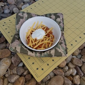 May include: A white bowl filled with chili, topped with shredded cheddar cheese and a dollop of sour cream, sits on a camouflage-patterned hot pad. The hot pad rests on a yellow cutting mat, surrounded by smooth stones.