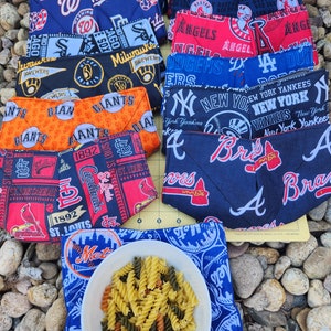 May include: A collection of fabric bowl cozies featuring various Major League Baseball team logos and colors. The cozies are arranged around a white bowl filled with tri-color rotini pasta. The cozies are made of quilted fabric.