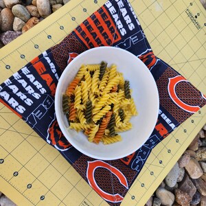 May include: A blue and orange fabric pot holder with the word "BEARS" repeated on it. A white bowl with cooked pasta is on top of the pot holder.