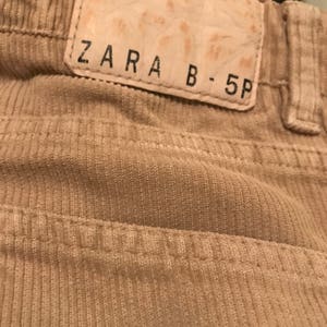 K&ouml;nnte beinhalten: Nahaufnahme eines beigefarbenen Cordstoffs mit einem braunen Lederflicken, auf dem "ZARA B-5P" steht.