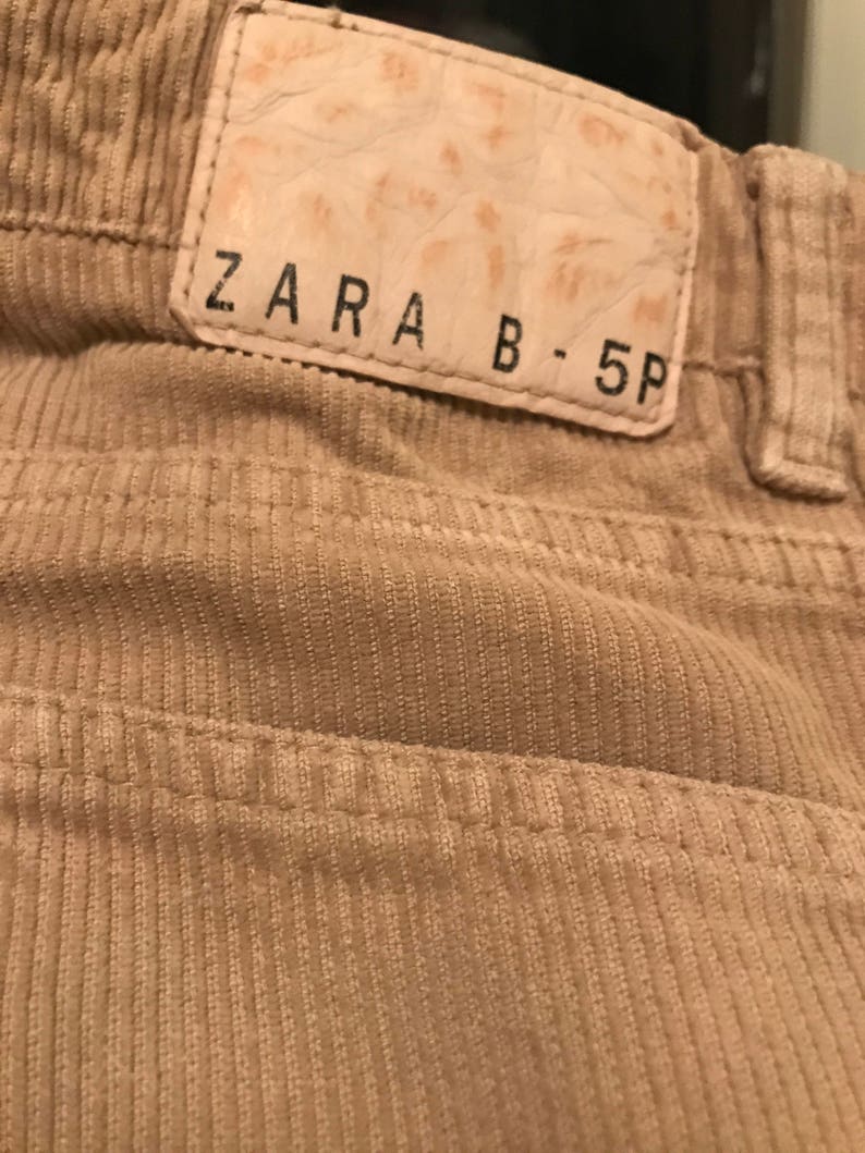 K&ouml;nnte beinhalten: Nahaufnahme eines beigefarbenen Cordstoffs mit einem braunen Lederflicken, auf dem "ZARA B-5P" steht.