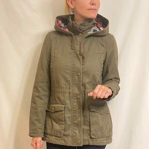 Op de afbeelding: Een groene capuchonjas met een geruite voering. De jas heeft een ritssluiting, een knoopsluiting en meerdere zakken.