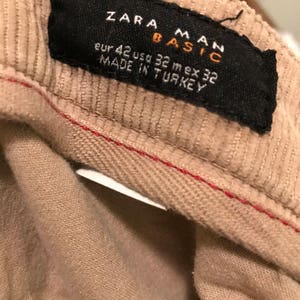 K&ouml;nnte beinhalten: Ein beigefarbenes Cordstoff-Etikett mit schwarzem Text, der "Zara Man Basic" und "Eur 42 Usd 32 Mex 32 Made in Turkey" lautet.