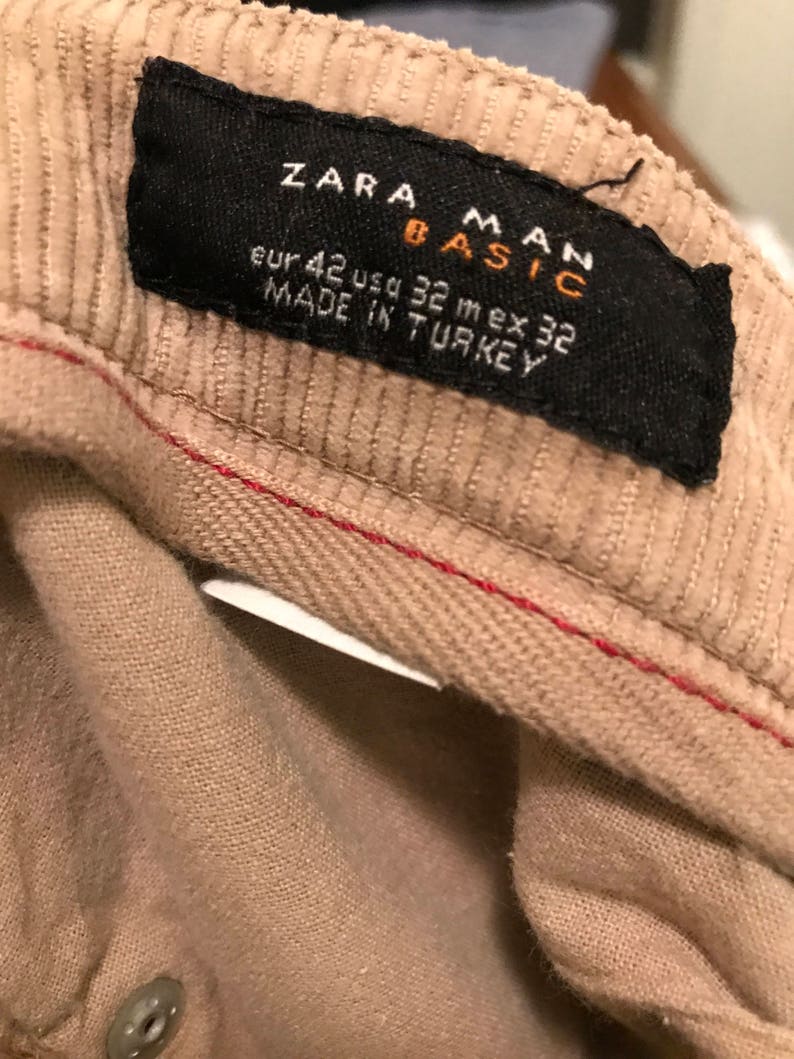 K&ouml;nnte beinhalten: Ein beigefarbenes Cordstoff-Etikett mit schwarzem Text, der "Zara Man Basic" und "Eur 42 Usd 32 Mex 32 Made in Turkey" lautet.