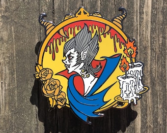 May include: A colorful enamel pin featuring a vampire with fangs, wearing a blue cape and holding a candle. The pin is set against a yellow background with dripping red paint and roses.