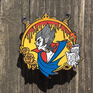 May include: A colorful enamel pin featuring a vampire with fangs, wearing a blue cape and holding a candle. The pin is set against a yellow background with dripping red paint and roses.