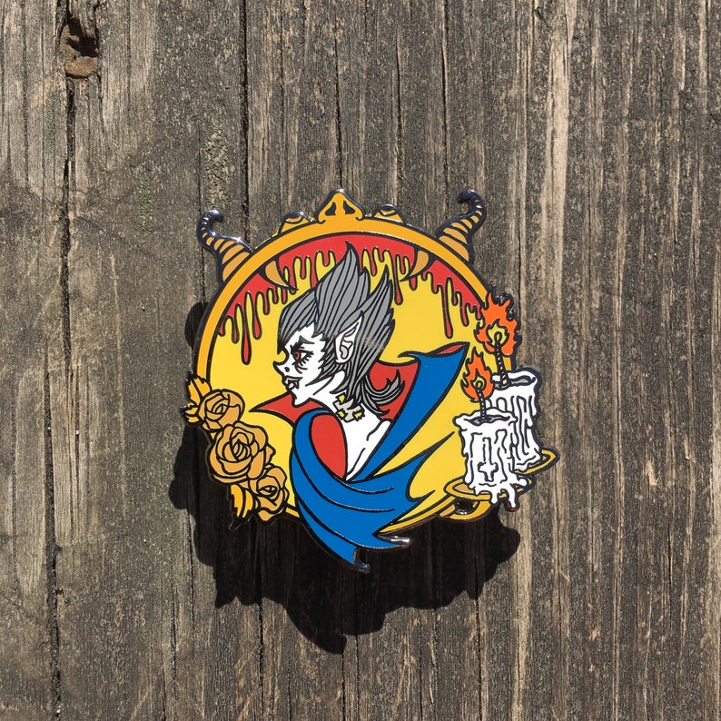 May include: A colorful enamel pin featuring a vampire with fangs, wearing a blue cape and holding a candle. The pin is set against a yellow background with dripping red paint and roses.