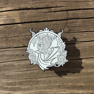 May include: White enamel pin featuring a black and white illustration of a creature with wings and horns, holding a candle. The creature is surrounded by dripping wax and roses.