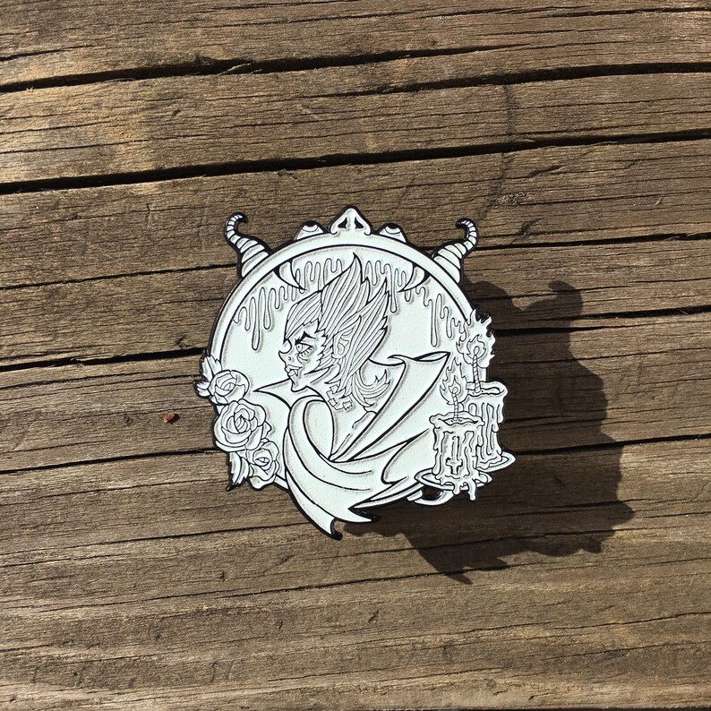 May include: White enamel pin featuring a black and white illustration of a creature with wings and horns, holding a candle. The creature is surrounded by dripping wax and roses.