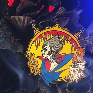 May include: A yellow, red, and blue enamel pin featuring a vampire with a candle and roses. The vampire has a pointed ear and is wearing a blue cape. The candle is lit and has a yellow flame.