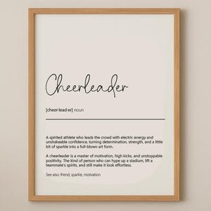 May include: A framed print featuring the word "Cheerleader" in cursive script. The definition describes a spirited athlete with electric energy and unshakeable confidence. The print is in a light wood frame.