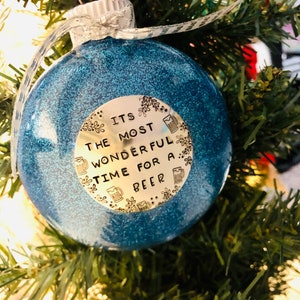 May include: A blue glitter Christmas ornament with a gold circle sticker that says "It's the most wonderful time for a beer".