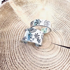 May include: Two silver metal bracelets with a white background and black floral and leaf designs. The bracelets are stacked on top of each other on a wooden surface.