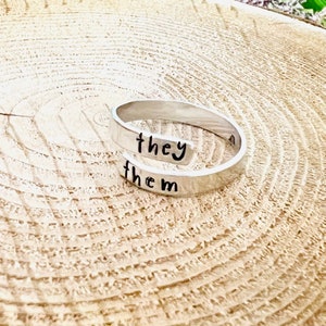 May include: A silver ring with the words "they them" engraved on it.
