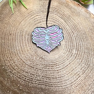 May include: A pink and blue enamel pin shaped like a heart with a ribcage design. The heart has a pink center and is dripping with pink and blue enamel.