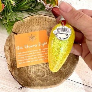May include: A yellow glitter-filled light bulb with a silver school bus charm that reads "School Bus" and "Ms. Peterson". An orange card with the text "Bus Drivers Light" is also present.
