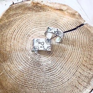 May include: Two silver metal bracelets with a white background and black floral and bone designs.