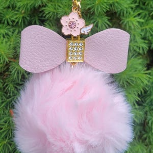 May include: A pink keychain with a fluffy pom-pom, a bow, and a flower charm. The flower charm has a pink center and white wings. The bow is made of pink leather. The keychain is hanging against a green background.