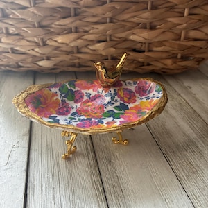 May include: An oval-shaped decorative dish featuring a vibrant floral pattern with pink, orange, and blue flowers on a white background. The dish has a gold-painted rim, three gold-toned legs, and a small gold bird figurine attached to the edge.