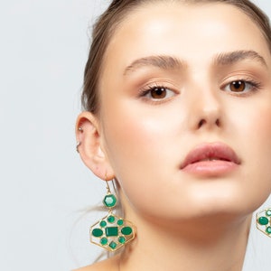 May include: A woman wearing gold earrings with green gemstone accents. The earrings are geometric in shape and feature multiple stones.