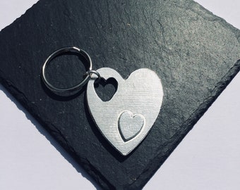 May include: Silver keychain with two interlocking heart shapes. The larger heart has a smaller heart cut out of it.