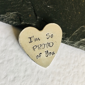 May include: A silver heart-shaped token with the words "I'm so proud of you" engraved on it.