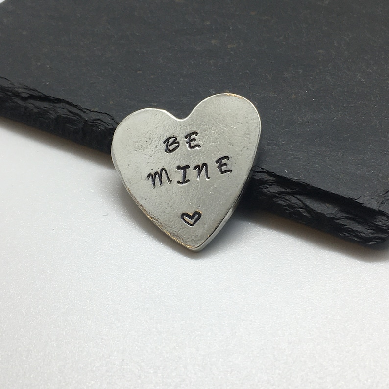May include: A silver heart-shaped charm with the words "BE MINE" engraved on it. A small heart is also engraved below the words.