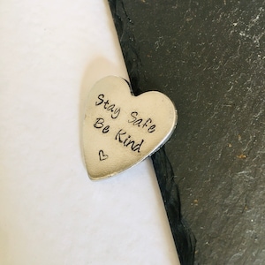 Può includere: Un ciondolo a forma di cuore argentato con le parole "Stay Safe Be Kind" incise su di esso.