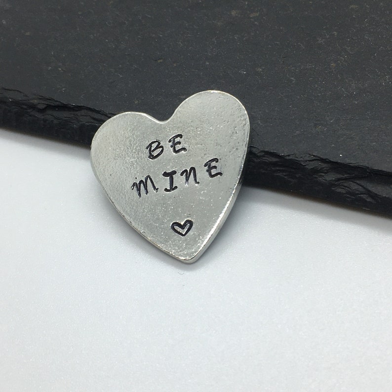 May include: A silver heart-shaped charm with the words "BE MINE" engraved on it. A small heart symbol is also engraved on the charm.