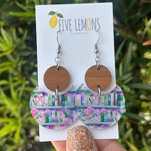 May include: A pair of dangle earrings featuring a wooden circle top and a semi-circle bottom with a floral and bookshelf design. The earrings are held up by silver hooks. The Five Lemons Company logo is visible on the white card.