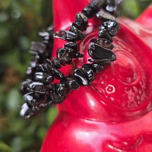 May include: A black gemstone bracelet made of polished, irregularly shaped stones. The bracelet is set against a bright red background, with a blurred green backdrop suggesting an outdoor setting. The stones appear to be obsidian.
