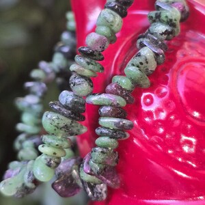 May include: Close-up of two strands of gemstone beads. The beads are a mix of green, black, and maroon colours. The beads are strung together, creating a necklace or bracelet. The background is a blurred red object.