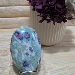 May include: An oval-shaped, polished stone in shades of teal, magenta, and white, displayed on a clear acrylic stand. The stone's surface features a unique pattern. A white vase with purple flowers is in the background. The stone is approximately 10 cm in height.