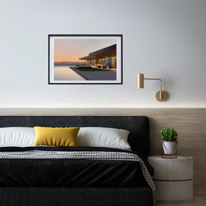 May include: A framed photograph of a modern house with a swimming pool and a view of a sunset. The house has large windows and a patio with chairs. The photo is hanging on a white wall above a black headboard.