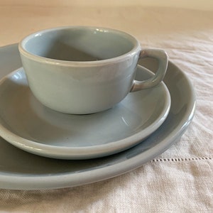May include: A light blue ceramic teacup with a matching saucer and a larger plate. The cup has a square handle. The set is a soft, muted blue color, and the pieces are stacked on top of each other.